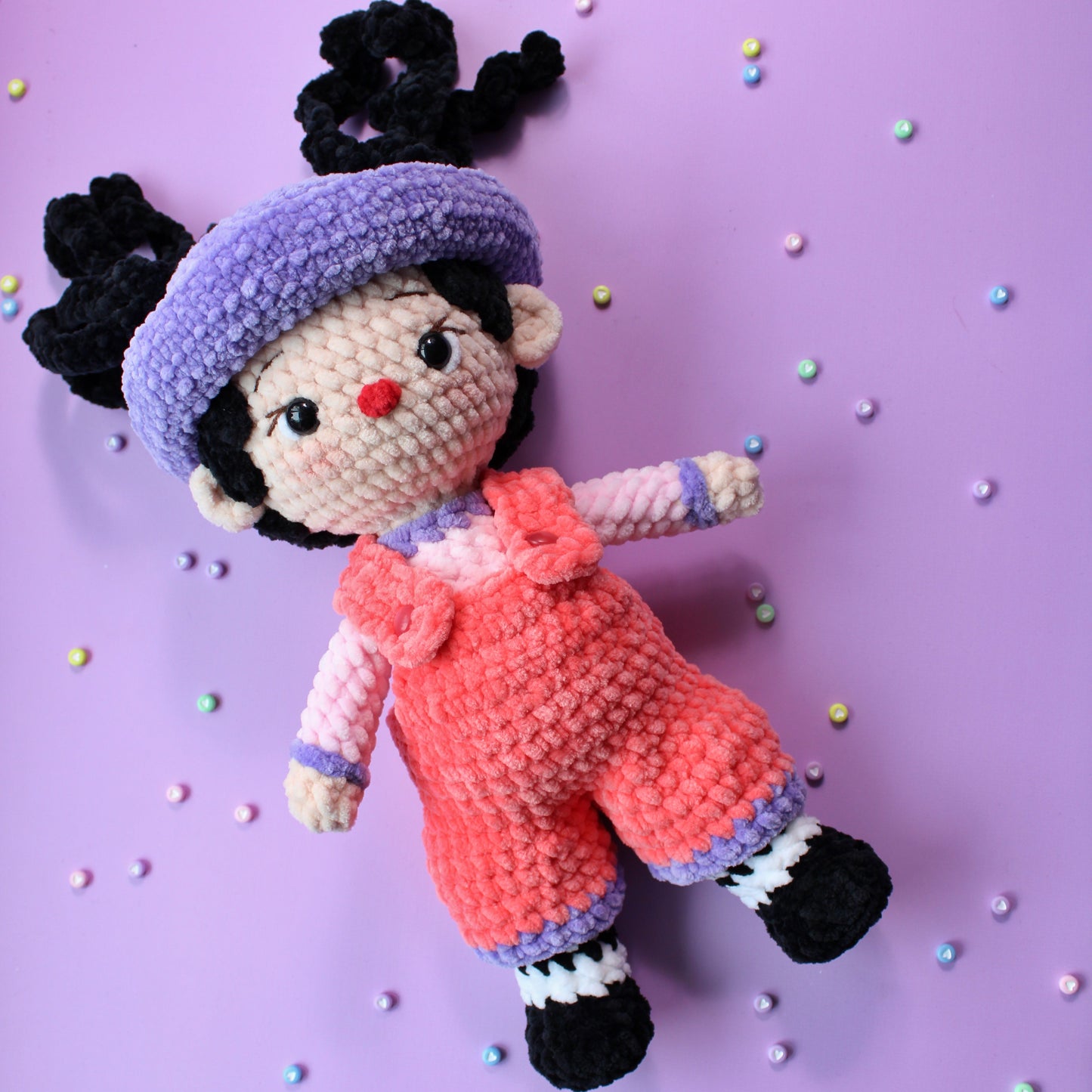 Crocheted doll with a purple hat and pink overalls on a purple background with colorful dots.
