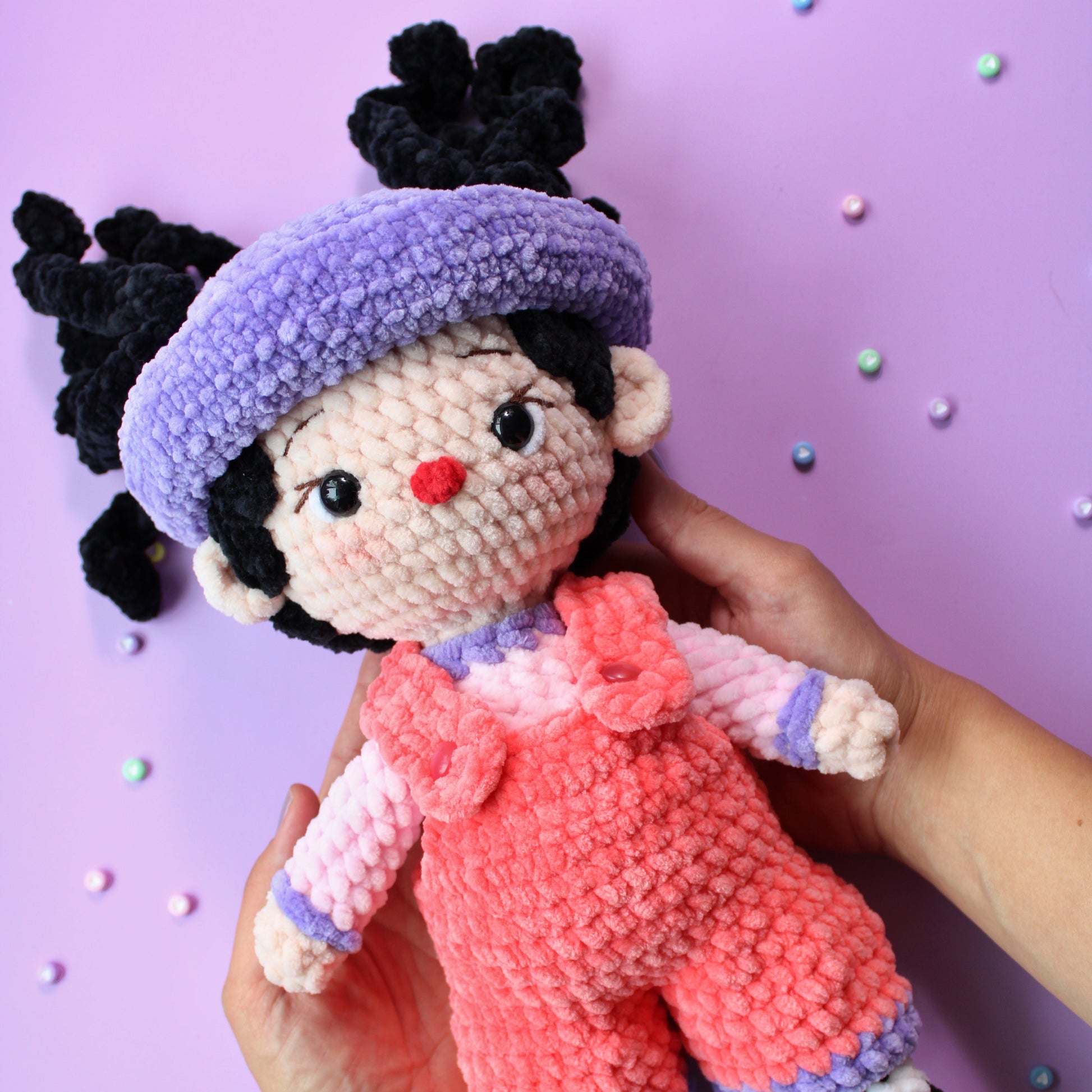 Hand holding a small crochet doll, Loonette the Clown, with pink overalls and black hair with a purple hat