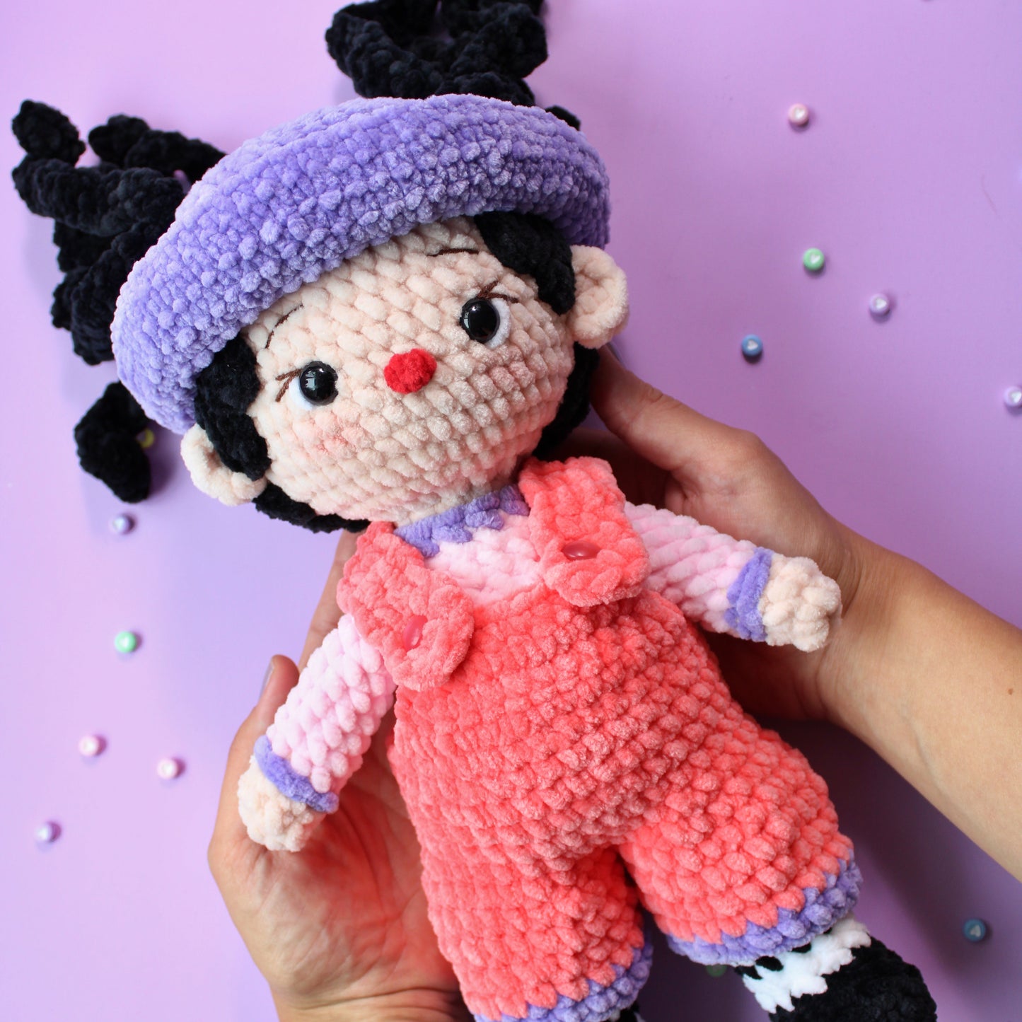 Hand holding a small crochet doll with pink overalls and a purple hat on a purple background.