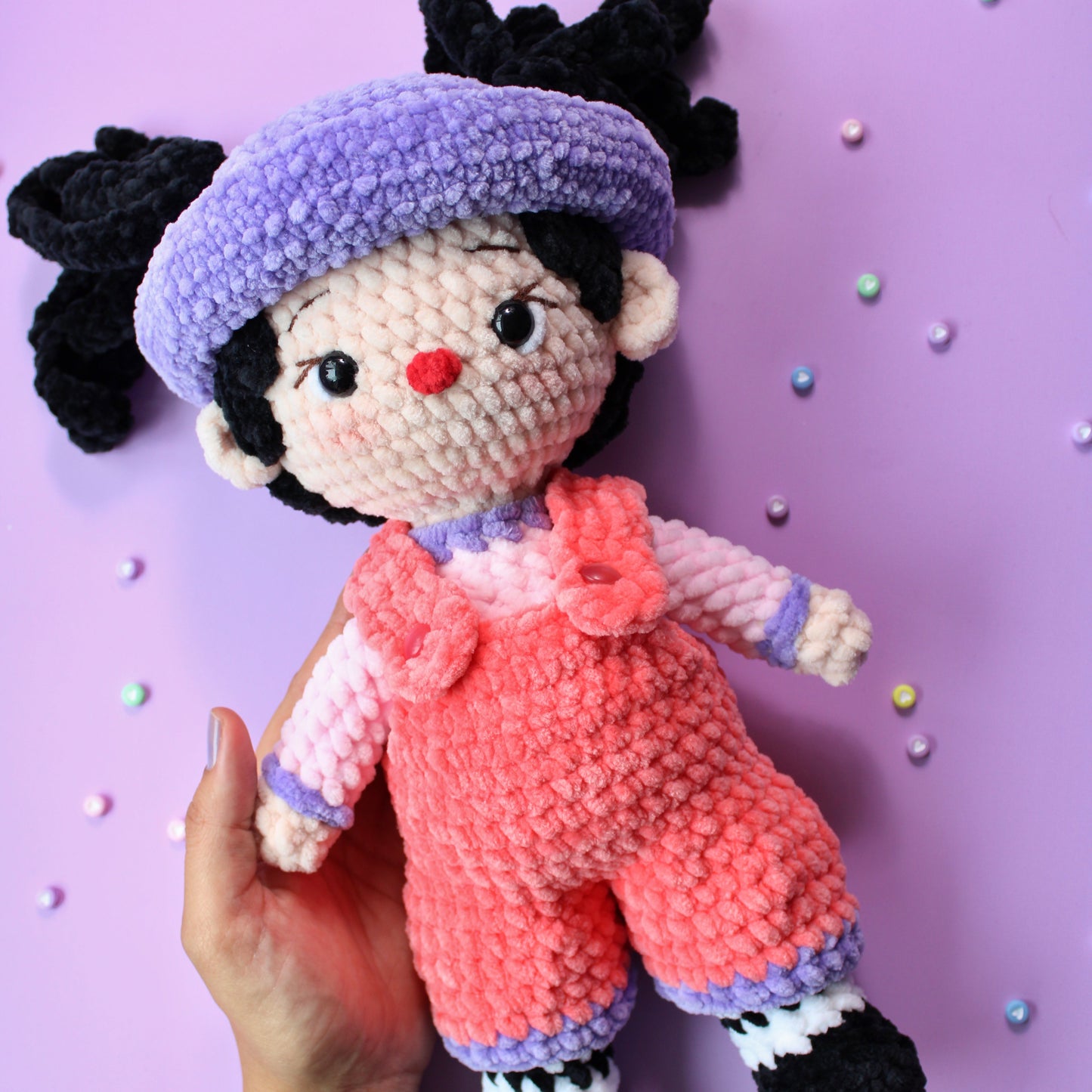 Handmade crochet doll with a purple hat and pink overalls on a purple background
